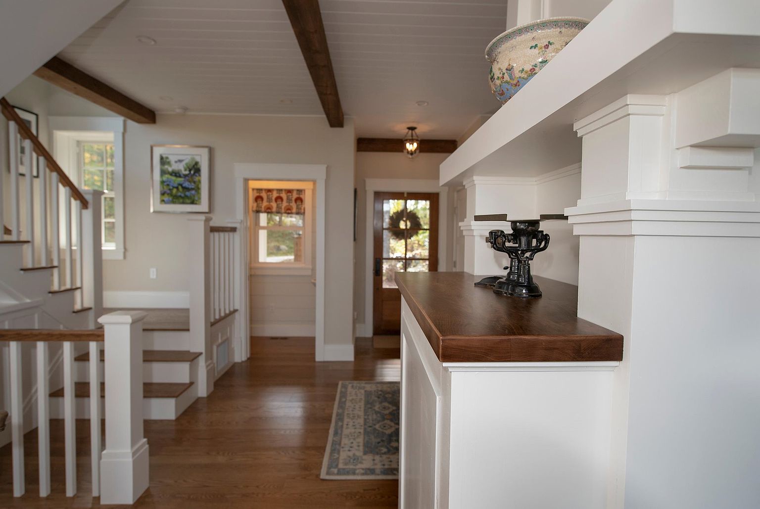 This interior view captures a bright, welcoming entryway and hallway featuring warm wood flooring and white trim. A staircase with a white railing ascends to the left, while a wooden console table with a decorative black scale sits in the foreground. The perspective looks through the space toward a front door with a wreath, highlighting the open, airy layout and traditional architectural details.