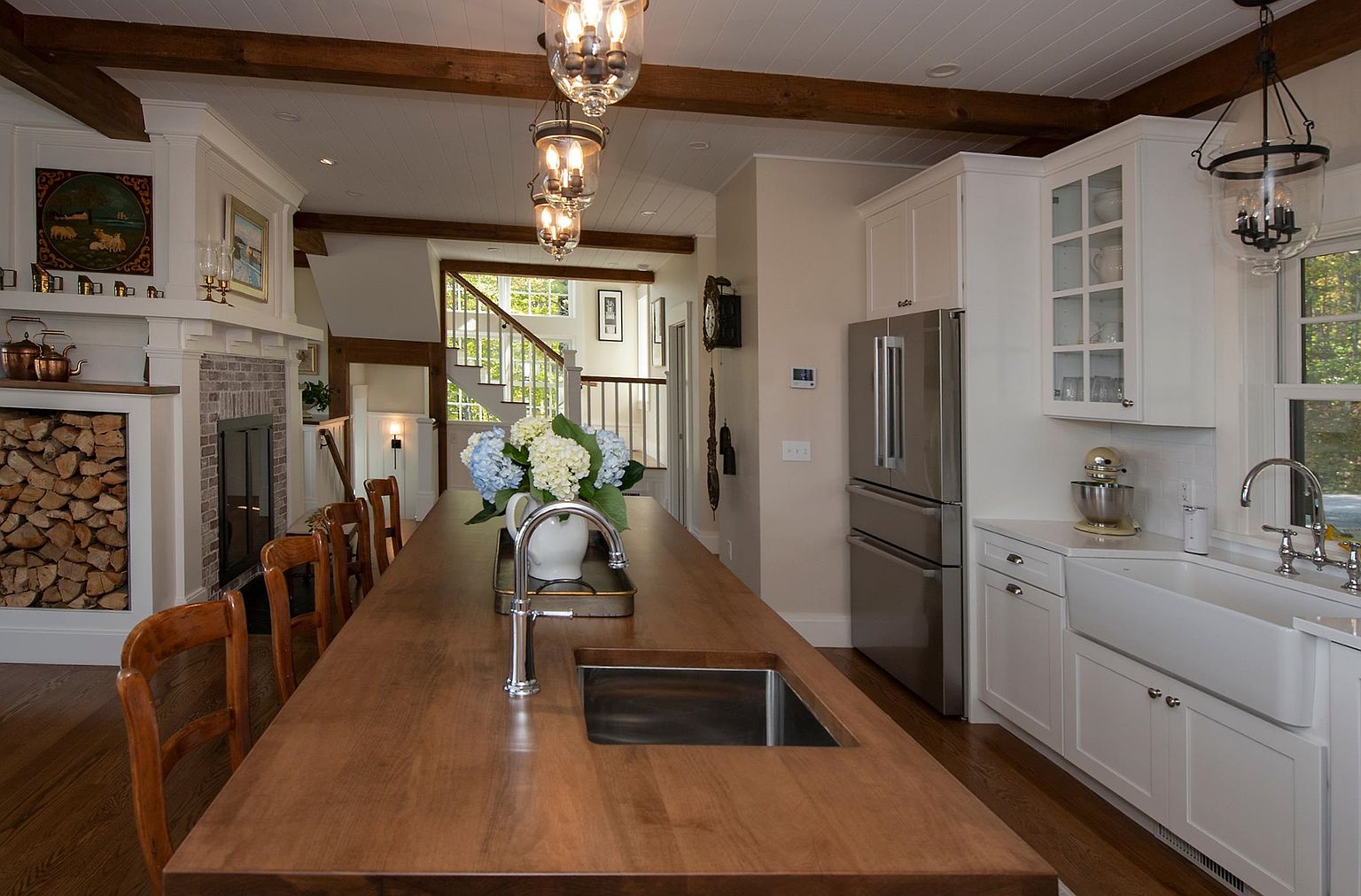 This inviting kitchen features a large central island with a wood countertop and a built-in sink, serving as the focal point of the open-concept space. White cabinetry and a farmhouse-style sink provide a classic aesthetic, while exposed wood ceiling beams and a stone-clad chimney breast near the fireplace add rustic charm. The perspective looks across the island toward a staircase in the background, highlighting the home's warm, cohesive design.