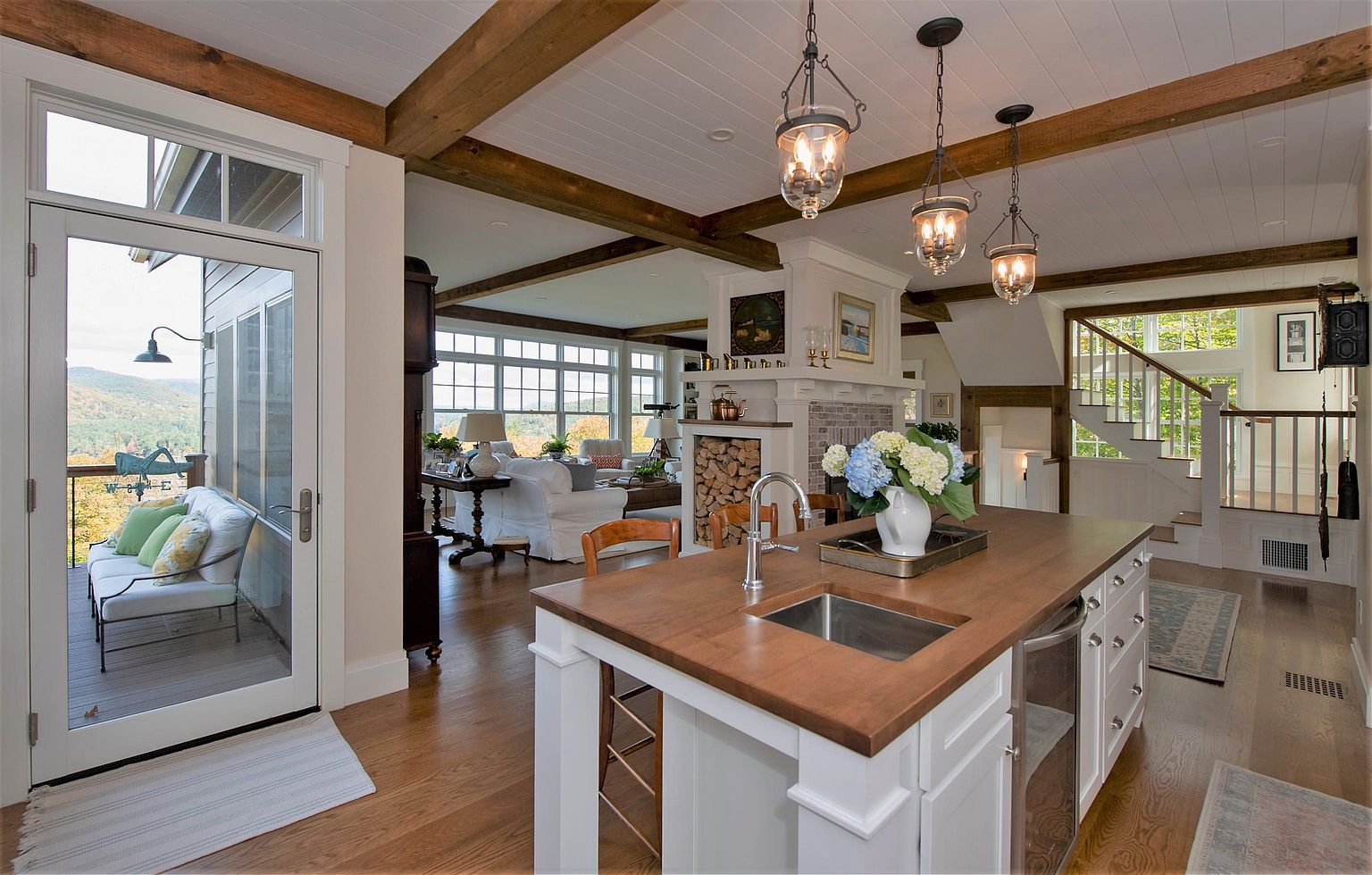 This open-concept kitchen features a large central island with a wood countertop and a built-in sink, serving as the focal point of the space. The room is characterized by exposed wooden ceiling beams, white cabinetry, and a seamless transition into an adjacent living area with a stone-clad fireplace and a view of a deck. The perspective captures the airy, inviting atmosphere of the home, highlighting the connection between the indoor living spaces and the scenic outdoor surroundings.