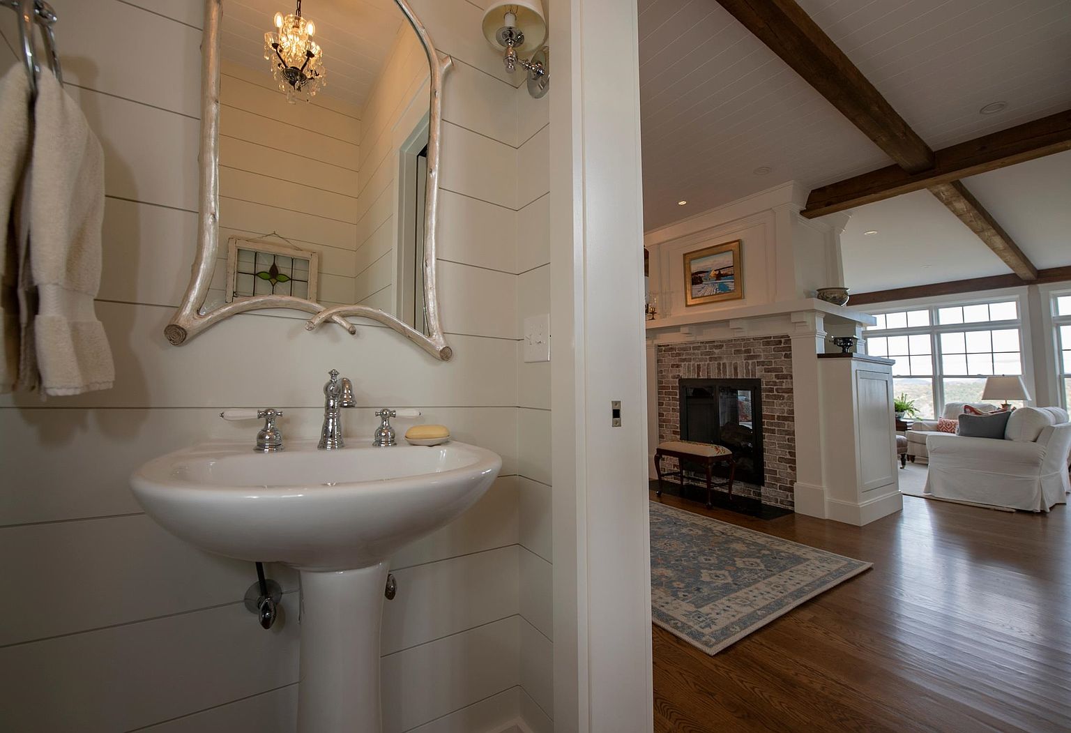 This image captures a view from a powder room looking out into an adjacent living area. The foreground features a pedestal sink with a unique, branch-like mirror frame and shiplap walls, while the background reveals a cozy living space with a brick-surround fireplace, exposed wooden ceiling beams, and comfortable white seating. The perspective creates a seamless transition between the intimate bathroom and the open-concept living area, highlighting the home's cohesive, rustic-inspired design.