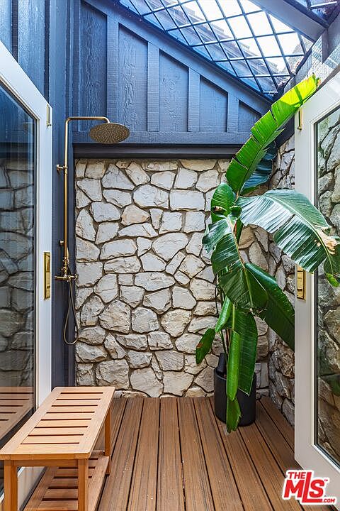 This image showcases an outdoor shower area, featuring a stone wall backdrop and a wooden deck. A brass shower fixture is mounted on the wall, and a large potted plant adds a touch of greenery. A wooden bench sits to the side, creating a spa-like atmosphere.