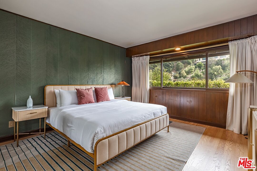 This is a primary bedroom featuring a king-sized bed with a beige upholstered frame and white linens, accented by two rose-colored pillows. The room has a unique design with a green textured wall behind the bed and wood-paneled walls with a large window offering a view of lush greenery. A modern nightstand with gold legs sits beside the bed, and a striped rug adds texture to the hardwood floor.