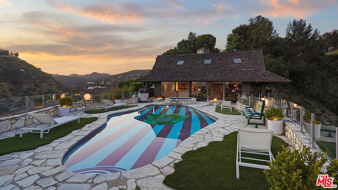 This image showcases a luxurious backyard with a uniquely designed pool featuring colorful stripes and a logo. The pool area is surrounded by stone paving, artificial turf, and lounge chairs, creating a relaxing outdoor space. The house, with its rustic roof and large windows, overlooks the pool and the scenic hillside beyond, offering a sense of privacy and tranquility.