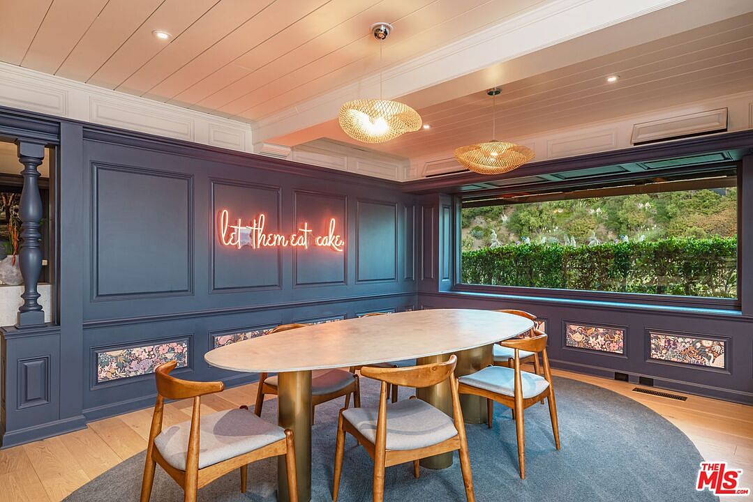 This is an interior shot of a dining room featuring a unique oval table with a light-colored top and gold cylindrical legs, surrounded by wooden chairs with light gray cushions. The room is decorated with dark blue paneled walls, a neon sign that reads "let them eat cake", and two modern, woven pendant lights hanging from the white, planked ceiling. A large window offers a view of lush greenery outside, and the room is grounded by a circular blue rug on the hardwood floor.