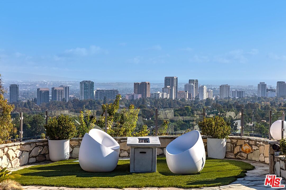 This exterior shot showcases a stylish patio or balcony area with a stunning city view. Two modern, egg-shaped chairs flank a square fire pit, creating an inviting outdoor living space. The area is bordered by a stone wall and glass railing, enhancing the view and adding a touch of elegance.