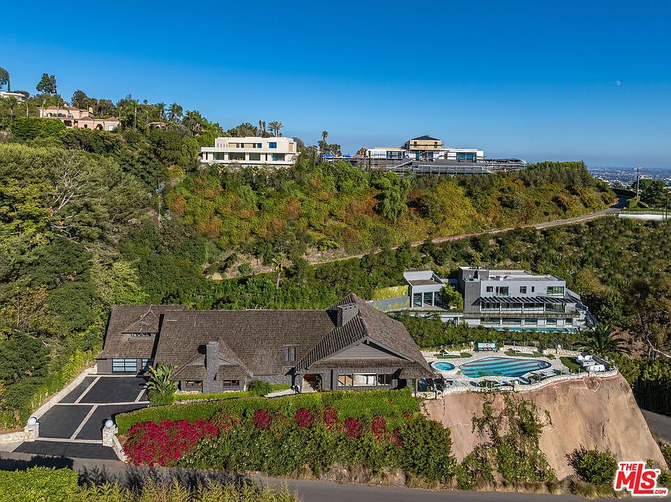 This aerial view showcases a luxurious estate nestled on a hillside. The property features a large, multi-level house with a dark roof, a modern pool area with a spa, and meticulously landscaped grounds. Several other modern homes are visible on the hillside, suggesting an exclusive neighborhood with stunning views.