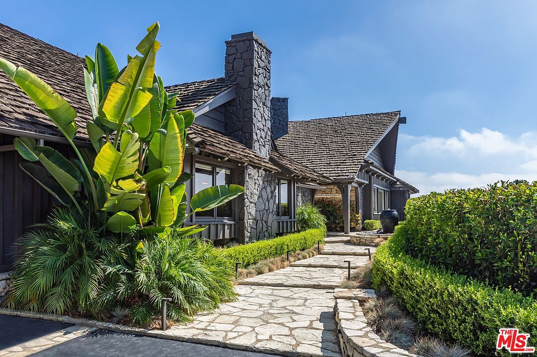 This is a front exterior view of a charming home with a stone pathway leading to the entrance. The house features a stone chimney, a wood shingle roof, and lush landscaping, including large tropical plants and manicured hedges. The overall impression is one of a well-maintained and inviting property.