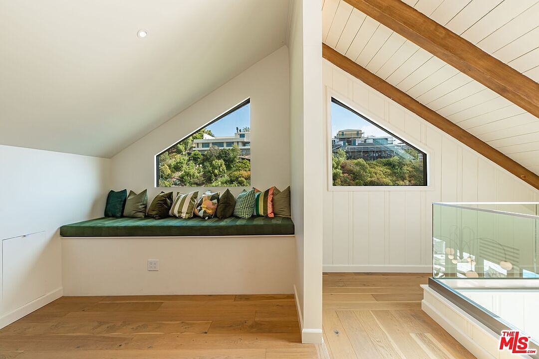 This interior shot showcases a cozy attic space featuring a built-in window seat with decorative pillows, offering a comfortable reading nook. The room is characterized by its angled ceilings with exposed wooden beams and white paneled walls, creating a bright and airy atmosphere. The hardwood flooring adds warmth and elegance to the space, enhancing its appeal as a unique and inviting area within the home.