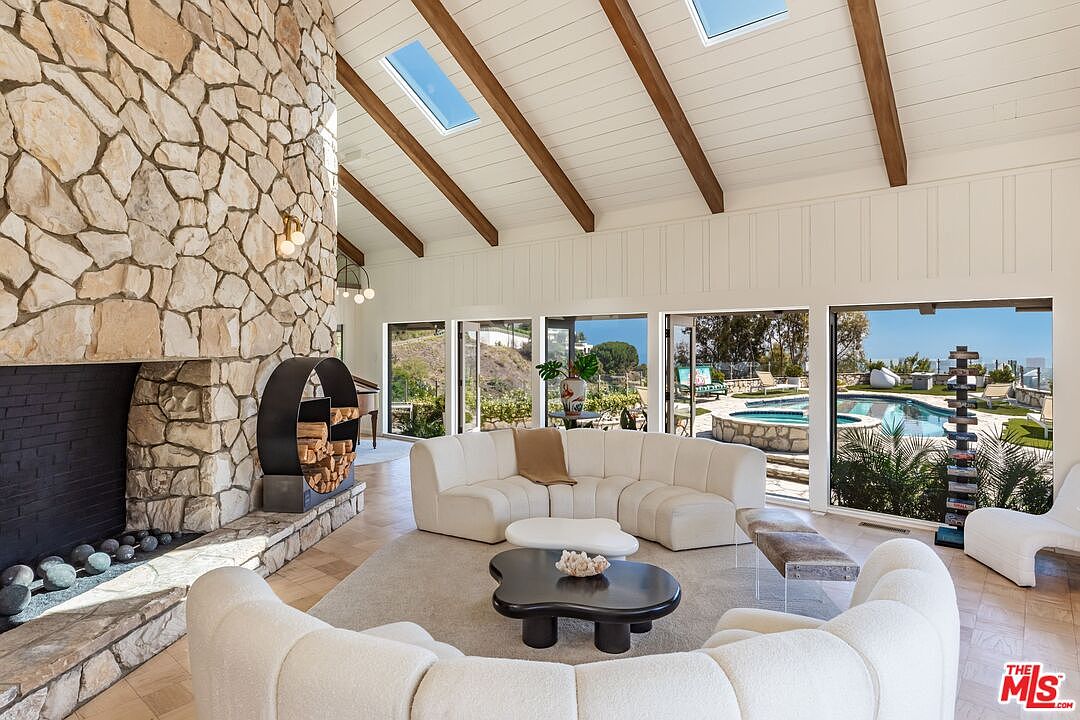 This is an interior shot of a living room featuring a stone fireplace, vaulted ceilings with exposed beams, and large windows overlooking a pool and landscaped yard. The room is furnished with a curved sofa and modern coffee tables, creating a bright and inviting space with a mid-century modern aesthetic. The perspective is from a low angle, emphasizing the height of the ceiling and the expansive view.
