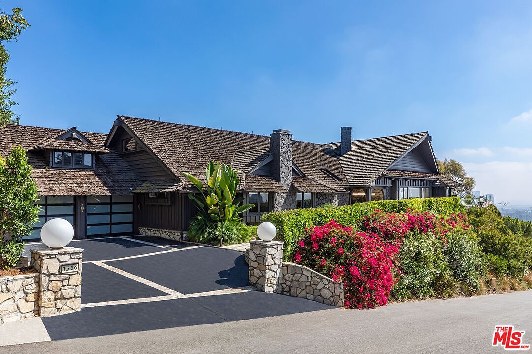 This is a front view of a luxurious, dark-stained wood house with a brown shingle roof. The property features a stone chimney, a two-car garage with glass doors, and a beautifully landscaped front yard with vibrant red bougainvillea and manicured hedges. A stone pillar with a spherical light fixture marks the entrance to the driveway, adding to the property's curb appeal.