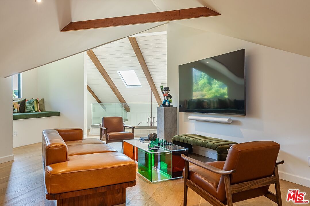 This interior shot showcases a stylish living room with a modern aesthetic. The room features a tan leather sectional sofa, two matching armchairs, and a unique glass coffee table with green and orange accents. A large flat-screen TV is mounted on the wall above a long, green velvet bench, and a decorative statue sits atop a gray pillar, adding a touch of personality to the space.