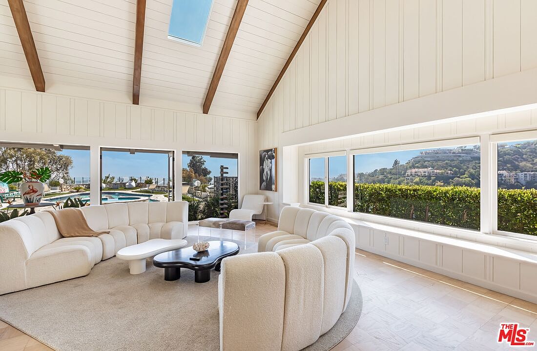 This is an interior shot of a bright and airy living room featuring vaulted ceilings with exposed wooden beams and white paneled walls. The room is furnished with two curved, cream-colored sofas, a unique coffee table, and a neutral-toned area rug. Large windows offer stunning views of the outdoors, including a pool and lush greenery, creating a seamless indoor-outdoor living experience.