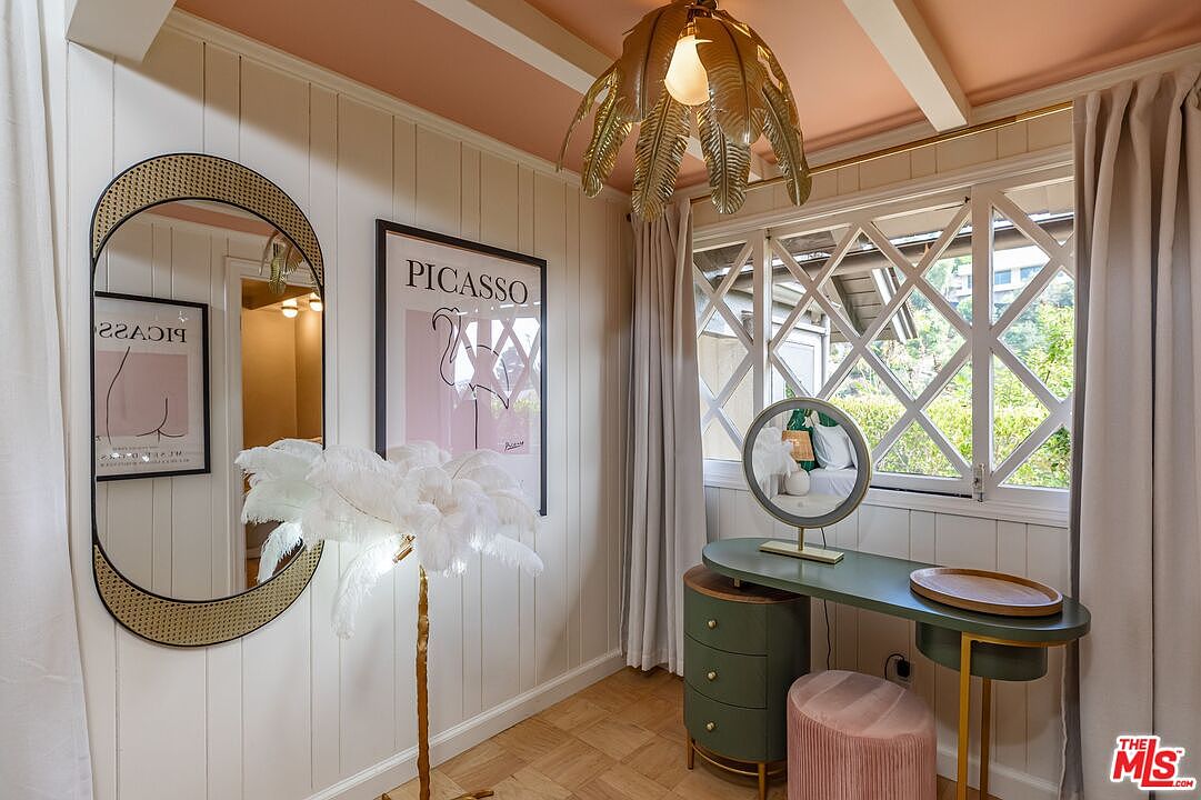 This is a charming bedroom featuring a vanity area with a green desk and a pink velvet stool. The room is decorated with a Picasso print, a unique palm-leaf chandelier, and a stylish mirror, creating a cozy and artistic atmosphere. The window provides natural light and a view of the outdoors.