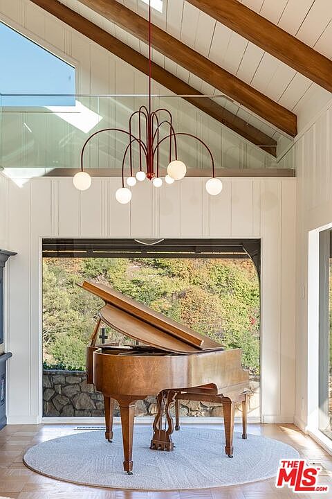 This interior shot showcases a grand living room featuring a grand piano as the focal point, positioned in front of a large window offering a view of lush greenery. A modern chandelier hangs above, complementing the room's high ceilings with exposed wooden beams. The space is bright and airy, creating an elegant and inviting atmosphere.