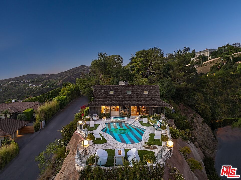 This aerial view showcases a stunning property perched on a hillside, featuring a unique pool with blue and black stripes surrounded by a stone patio and lounge chairs. The house has a rustic charm with a dark shingled roof and multiple windows, blending seamlessly with the lush greenery and surrounding landscape. The overall impression is one of luxury and privacy.