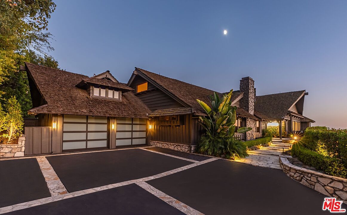 This is a front exterior view of a luxurious home at dusk, showcasing a well-maintained driveway with stone accents leading to a two-car garage. The house features a unique architectural style with a stone chimney, wood siding, and a shingled roof. Landscape lighting highlights the lush greenery and stone pathway leading to the entrance, creating a warm and inviting ambiance.