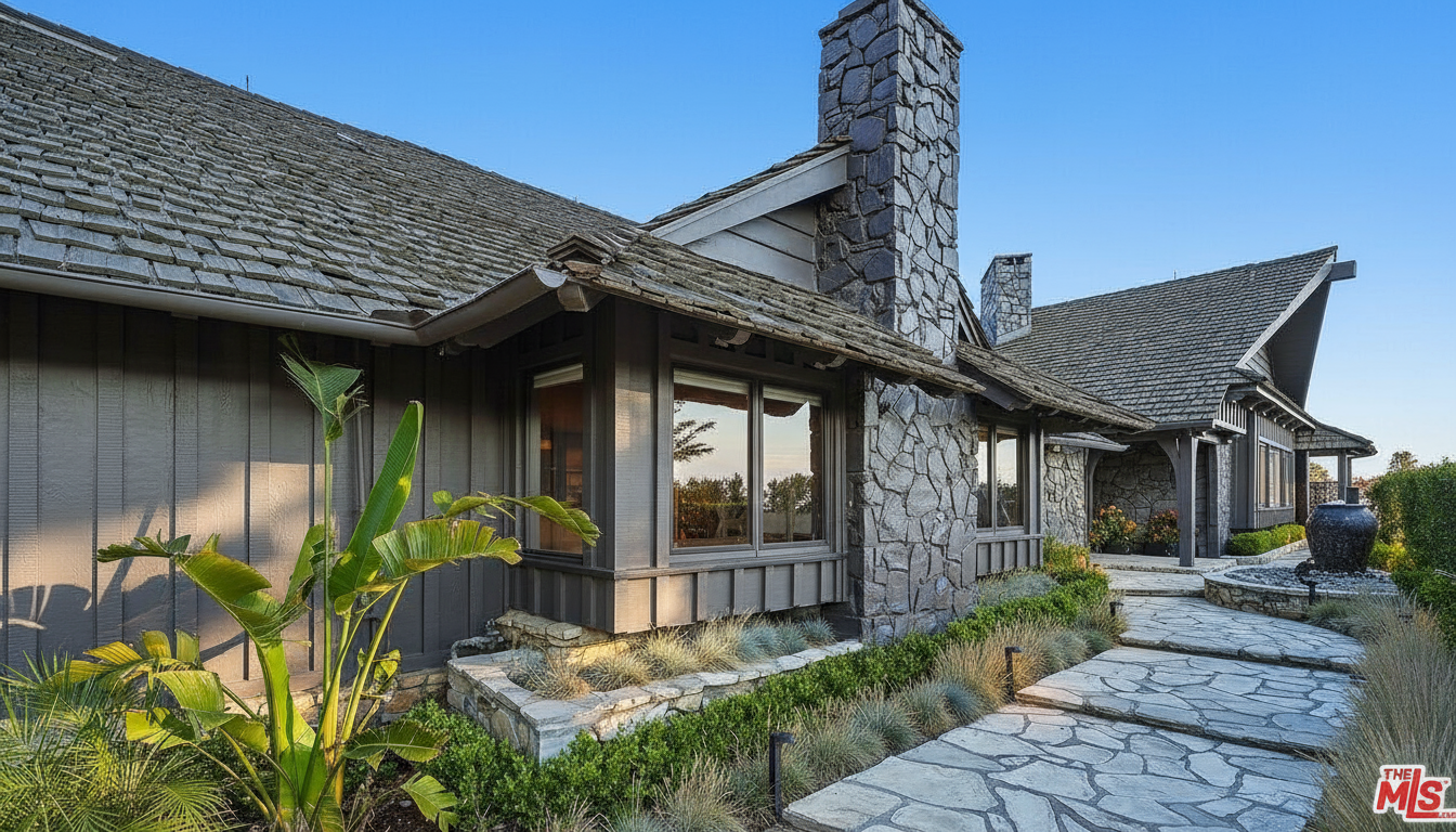 This is a side view of a house, showcasing its architectural design and exterior features. The building is stone and wood with a shingled roof and large chimneys. The landscape is well-manicured with stone walkways and various plants.