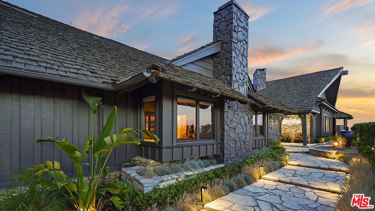 This image showcases the side exterior of a luxurious home, highlighting its stone chimney, wood siding, and shingle roof. A stone pathway leads towards the house, bordered by manicured landscaping and subtle lighting. The warm sunset in the background adds to the property's inviting and upscale ambiance.