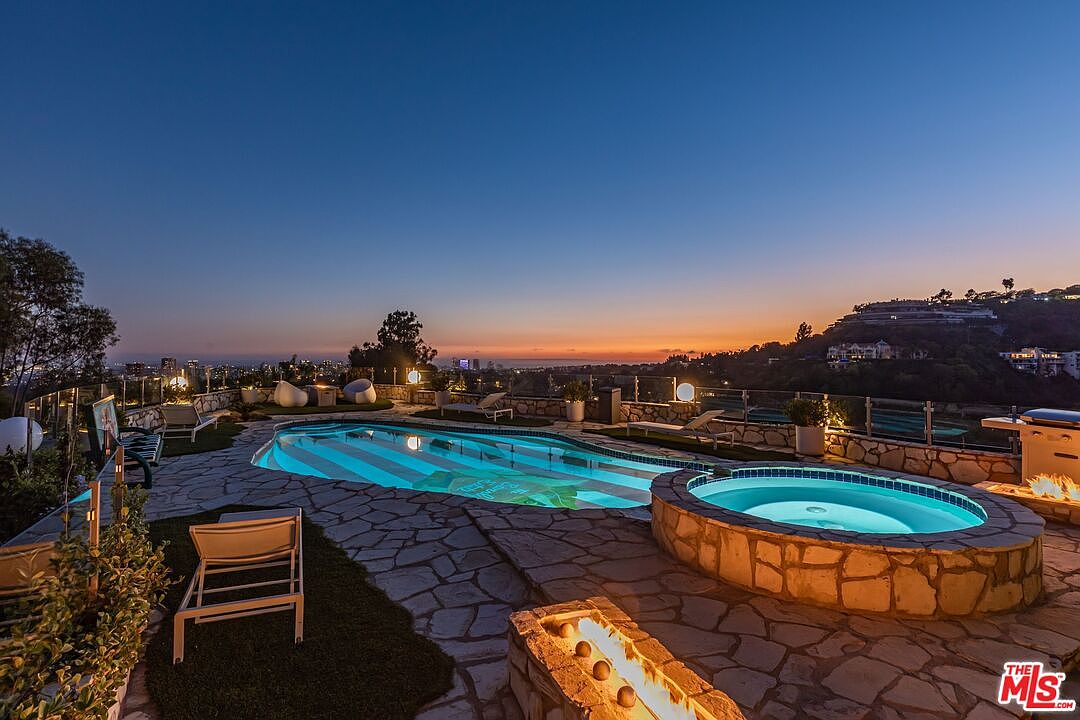 This image showcases a luxurious outdoor pool and spa area at dusk. The pool features a unique striped pattern, while the adjacent spa is constructed with natural stone. A fire pit adds warmth and ambiance, and the scene is set against a backdrop of city lights and a colorful sunset, creating an inviting and upscale atmosphere.