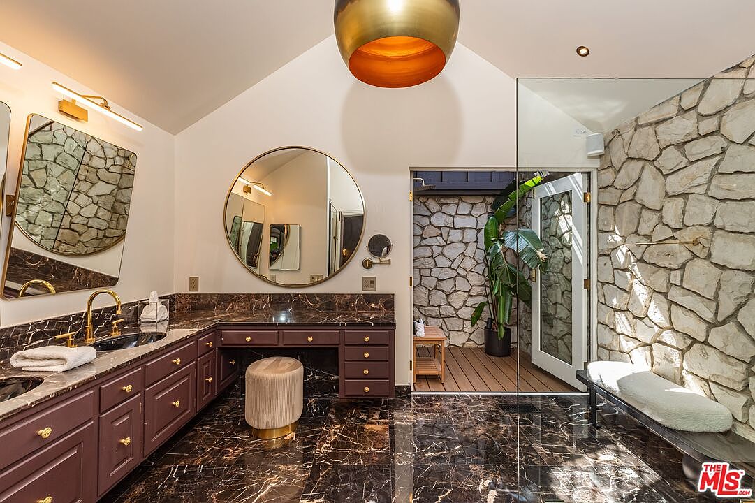 This primary bathroom features a unique design with a stone accent wall and a vanity area with a dark countertop and cabinetry. A round mirror hangs above the vanity, complemented by a modern light fixture. The flooring is a dark marble, adding a touch of luxury to the space, and a door leads to an outdoor area.