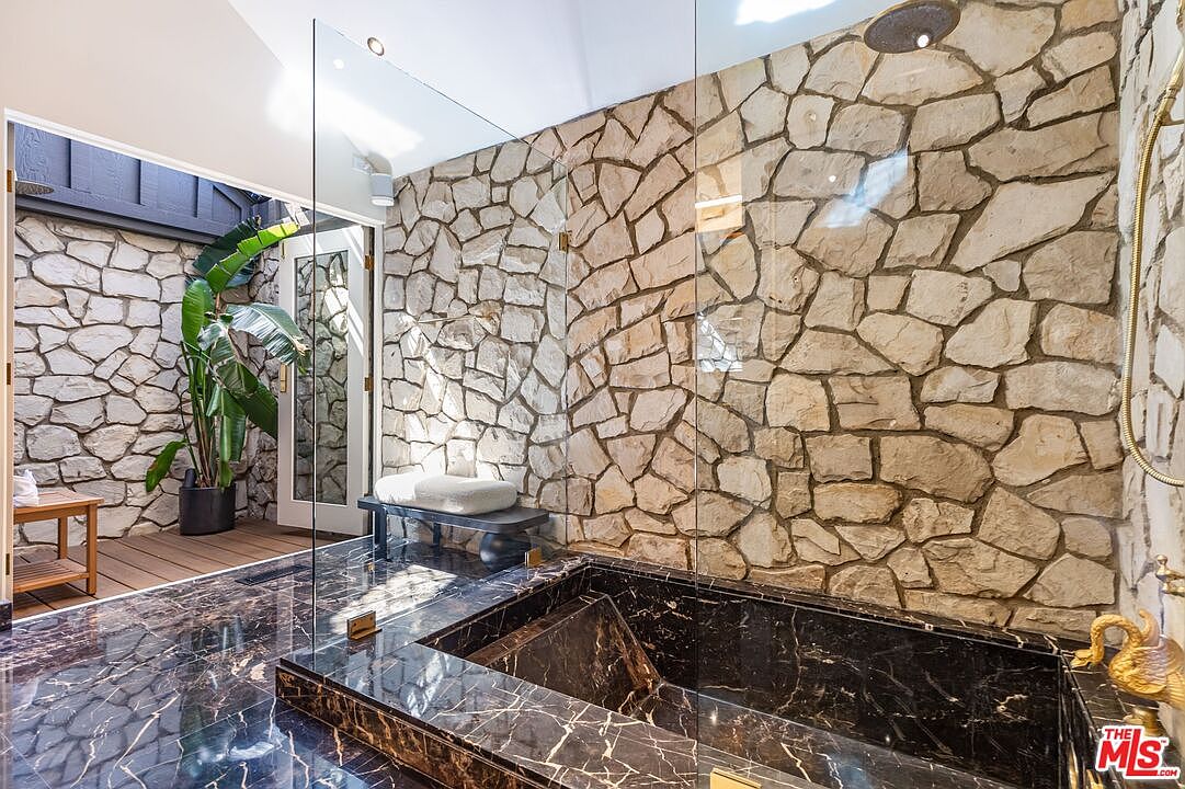 This primary bathroom features a luxurious black marble bathtub and flooring, complemented by a unique stone wall. A glass partition separates the bathing area, while a lush green plant adds a touch of nature. The overall design creates a spa-like atmosphere with a blend of modern and natural elements.