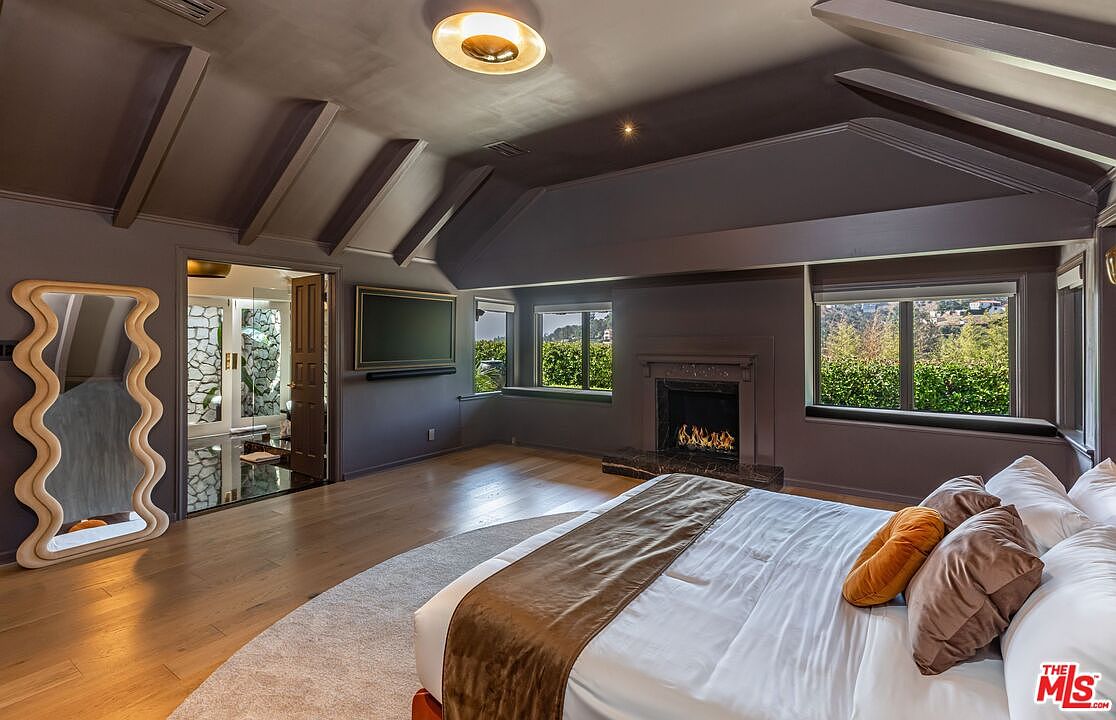 This is a primary bedroom featuring a king-size bed with white linens and brown and orange pillows, a fireplace, and two windows with views of greenery. The room has a dark gray color scheme with exposed beams on the ceiling and a unique wavy mirror. The flooring is light wood, and there is a doorway leading to another room, possibly a bathroom.