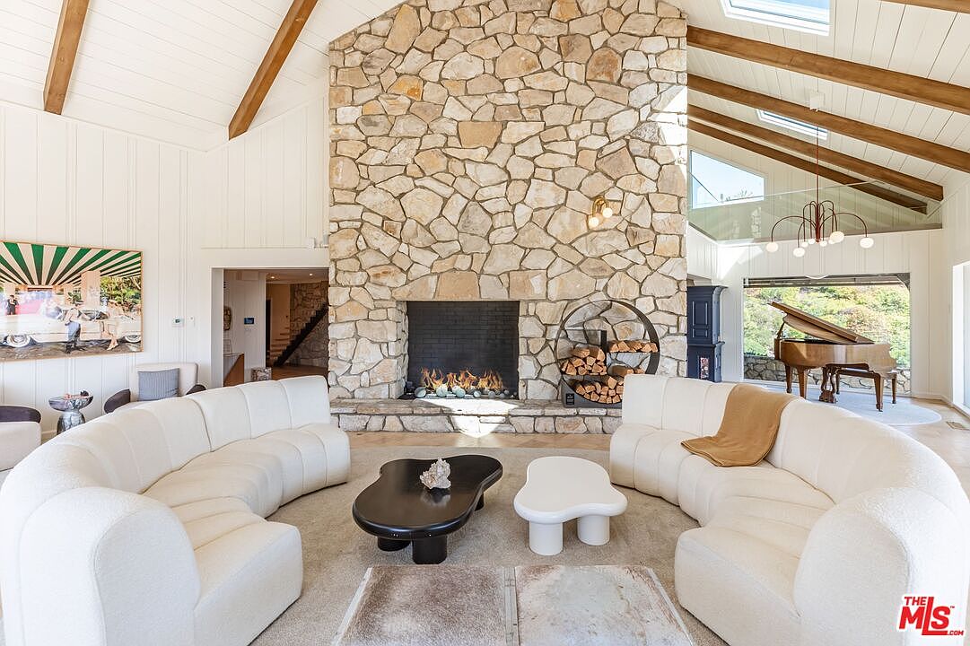 This is a spacious living room featuring a stone fireplace as a central focal point, flanked by curved white sofas. The room has high ceilings with exposed wooden beams and skylights, providing ample natural light. A grand piano sits near a large window, adding a touch of elegance to the space.