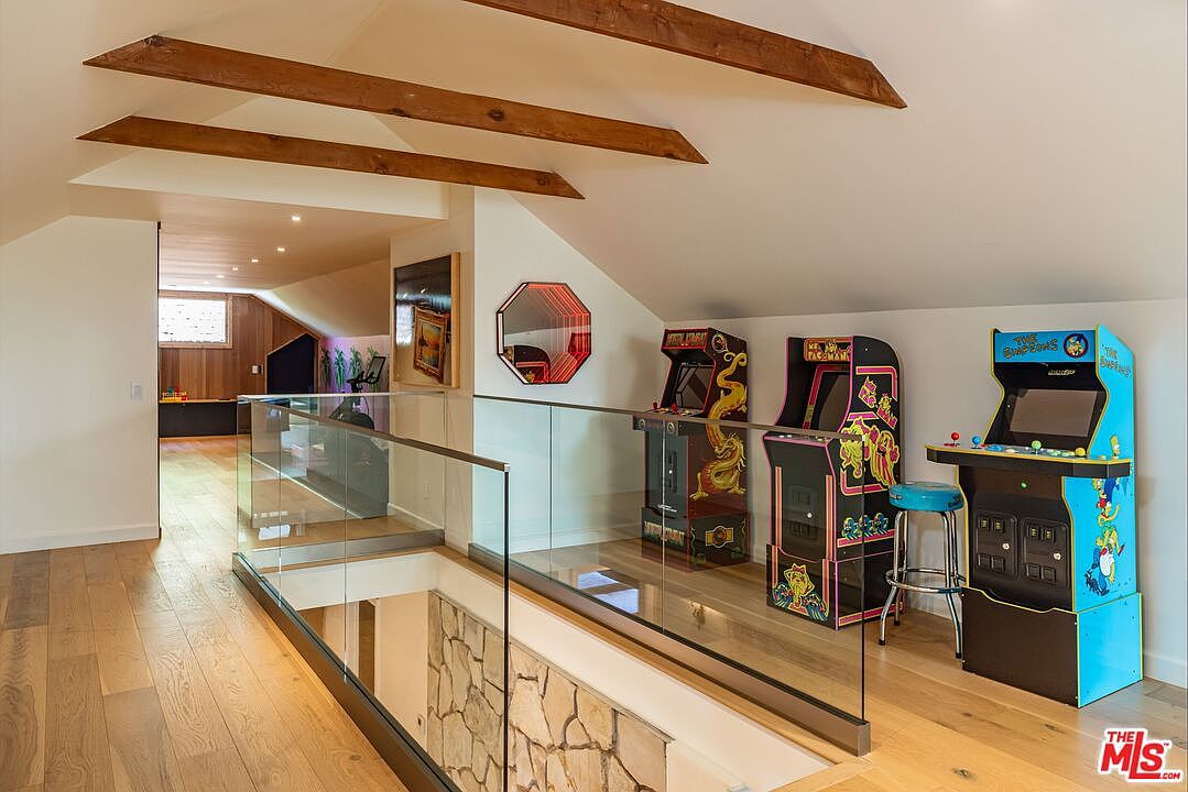 This interior shot showcases a hallway area with a unique design, featuring wooden beams on the ceiling and a glass-paneled railing overlooking a lower level with stone accents. Several arcade game machines are lined up against the wall, adding a playful touch to the space. The hardwood flooring and natural light create a warm and inviting atmosphere.