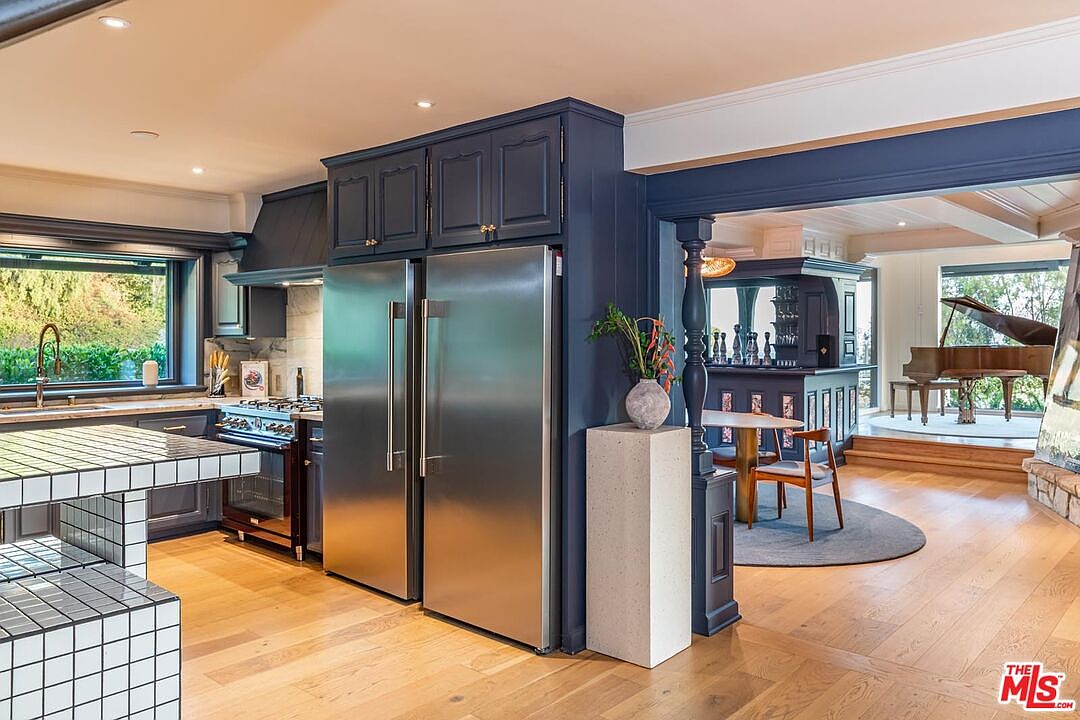 This interior shot showcases a well-appointed kitchen that seamlessly transitions into a living space with a bar and piano. The kitchen features dark blue cabinetry, stainless steel appliances, and a unique tiled island. The open layout and hardwood floors create a warm and inviting atmosphere, perfect for entertaining.