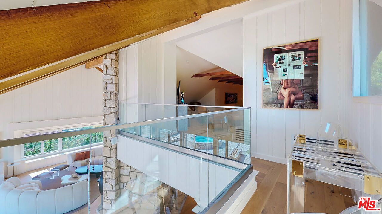 This interior shot showcases a modern hallway or landing area with a glass railing overlooking a living space below. The design features a blend of natural materials like wood and stone, complemented by white walls and contemporary artwork. The space is bright and airy, creating a sophisticated and inviting atmosphere.