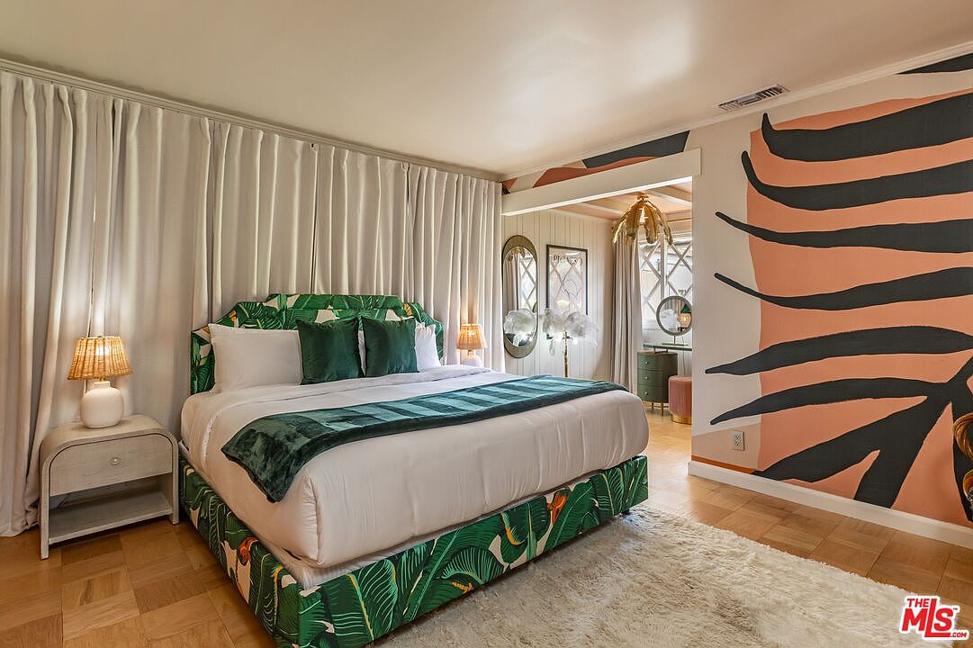 This is a stylish primary bedroom featuring a bed with a vibrant green leaf pattern on the headboard and base, complemented by green pillows and a throw. The room is decorated with a unique wall mural and soft, neutral curtains, creating a tropical yet modern aesthetic. A white nightstand with a lamp sits beside the bed, and a plush white rug adds texture to the wooden floor.