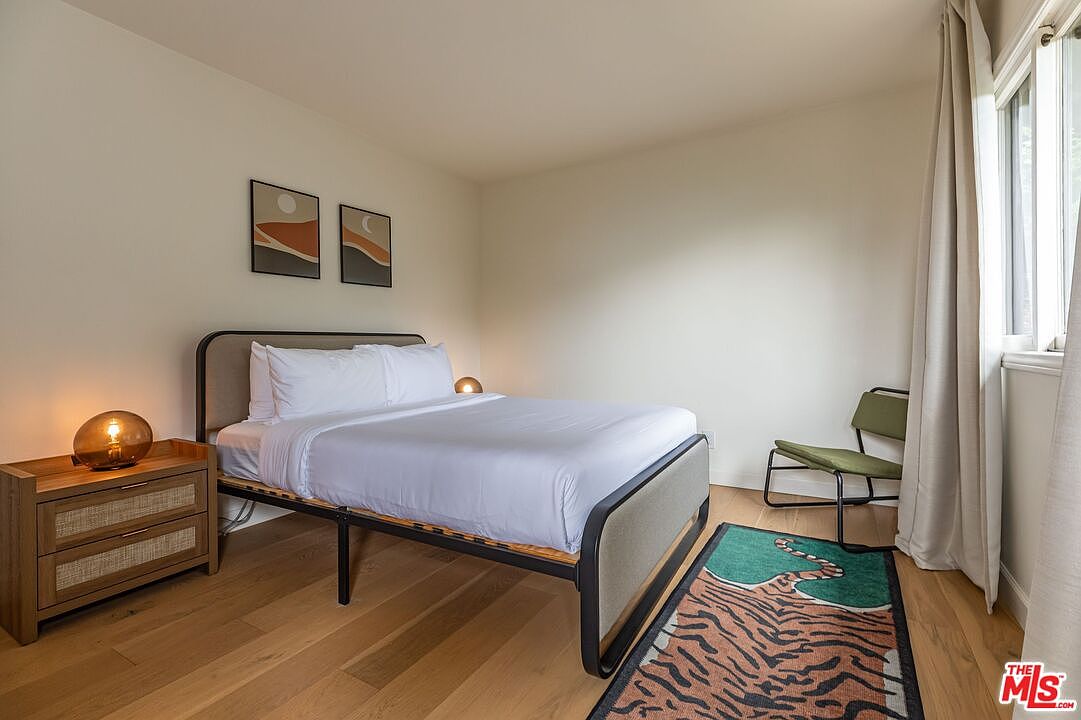 This is a cozy bedroom featuring a bed with white linens, a wooden nightstand with a lamp, and two framed artworks above the bed. A green chair sits near the window with beige curtains, and a tiger-striped rug adds a touch of character to the wooden floor. The room has a minimalist and inviting aesthetic.