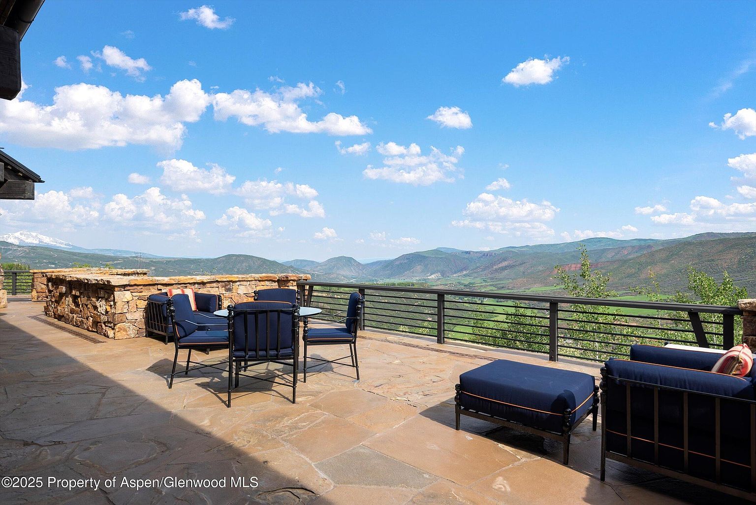 This image showcases a spacious patio or deck area with outdoor seating, offering stunning panoramic views of rolling hills and a clear blue sky with scattered clouds. The patio features stone flooring and a sturdy railing, creating an inviting outdoor living space perfect for relaxation and entertainment. The furniture is dark blue with a modern design.