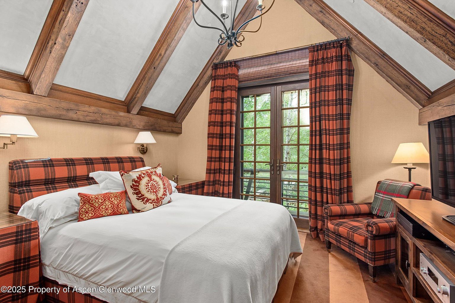 This is a cozy primary bedroom featuring a plaid upholstered bed frame and matching armchair, creating a warm and inviting atmosphere. The room has a vaulted ceiling with exposed wooden beams and a large window with plaid curtains, providing natural light and a view of the outdoors. The overall style is rustic and comfortable, perfect for a relaxing retreat.