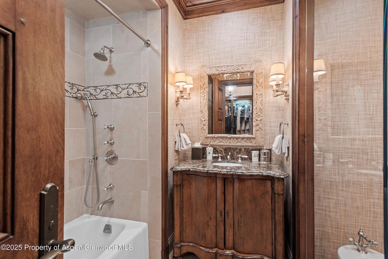 This is a well-appointed bathroom featuring a combination shower and tub with beige tiling and a decorative border. A wooden vanity with a marble countertop and an ornate mirror is the focal point, flanked by sconce lighting. The walls are covered in a textured wallpaper, creating a warm and inviting atmosphere.