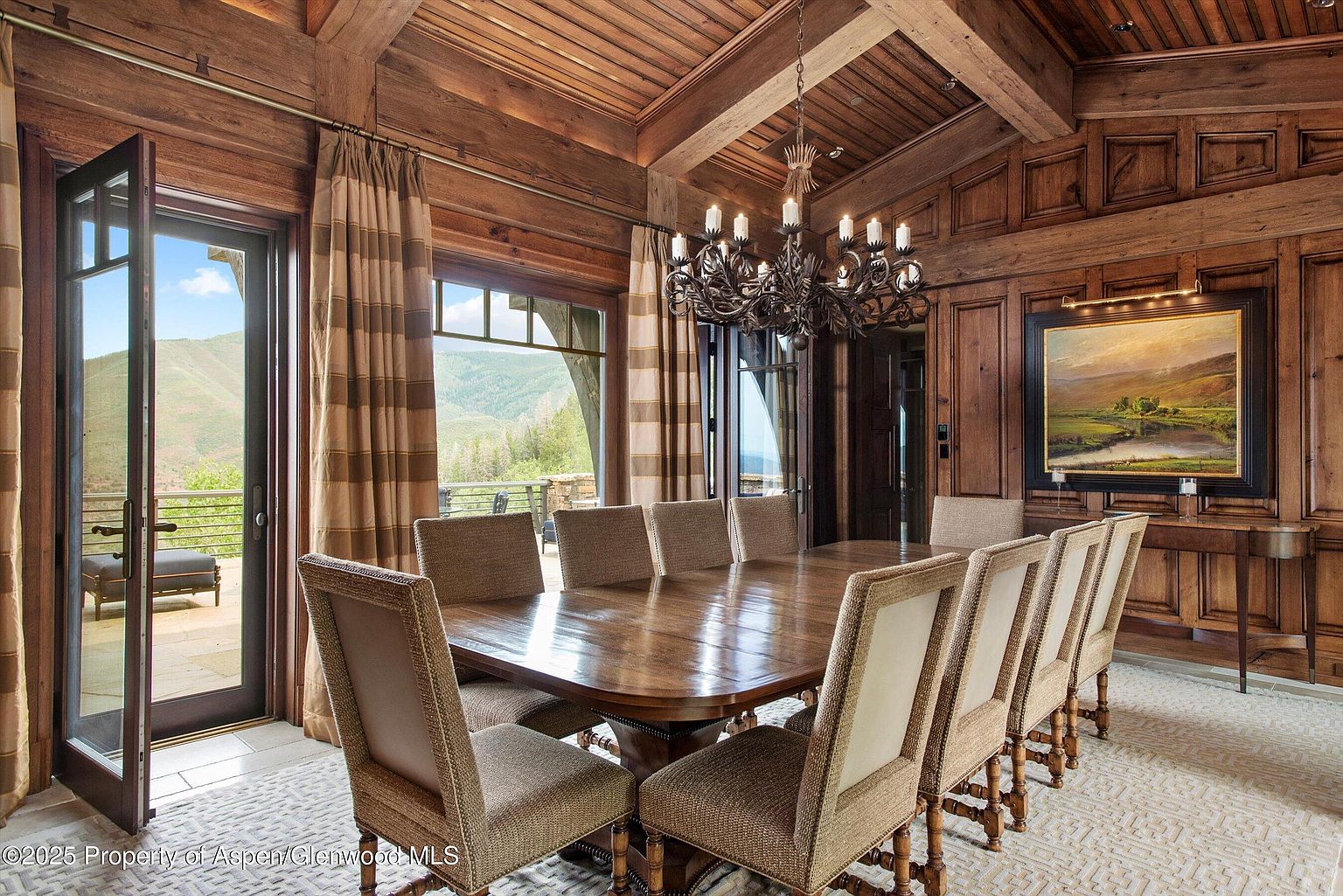 This is an elegant dining room featuring a large wooden table surrounded by upholstered chairs. The room is paneled with rich wood, complemented by a decorative chandelier and a landscape painting. Large windows and doors offer views of the outdoor scenery, creating a luxurious and inviting atmosphere.