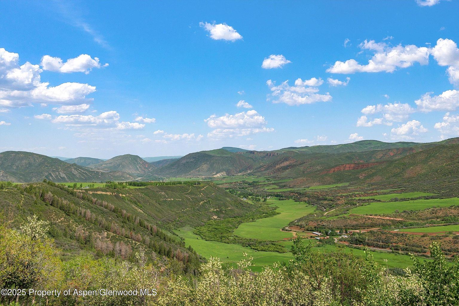 This aerial view showcases a sprawling green valley nestled between rolling hills and mountains under a bright blue sky dotted with fluffy white clouds. The landscape features a mix of lush meadows, forested areas, and winding waterways, creating a serene and picturesque setting. The perspective emphasizes the vastness and natural beauty of the property, highlighting its potential for a secluded and tranquil retreat.