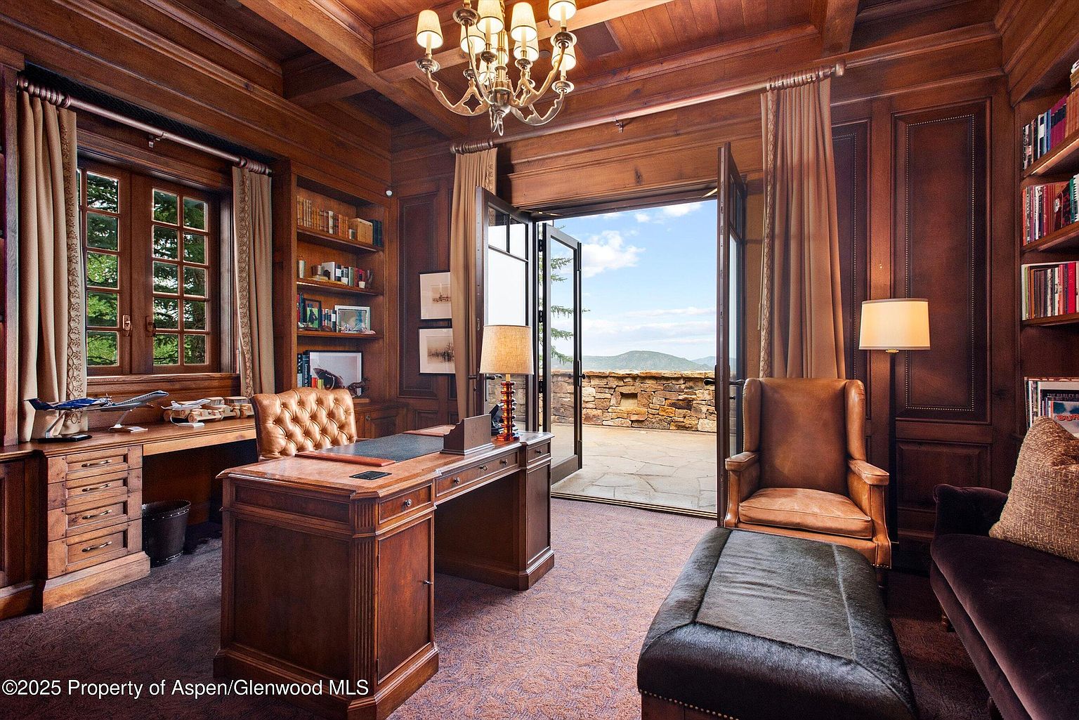 This is a luxurious home office featuring rich wood paneling, built-in bookshelves, and a large executive desk. The room opens onto a stone patio with a scenic mountain view, creating a sophisticated and inviting workspace. A leather armchair and ottoman provide a comfortable seating area, enhancing the room's appeal.