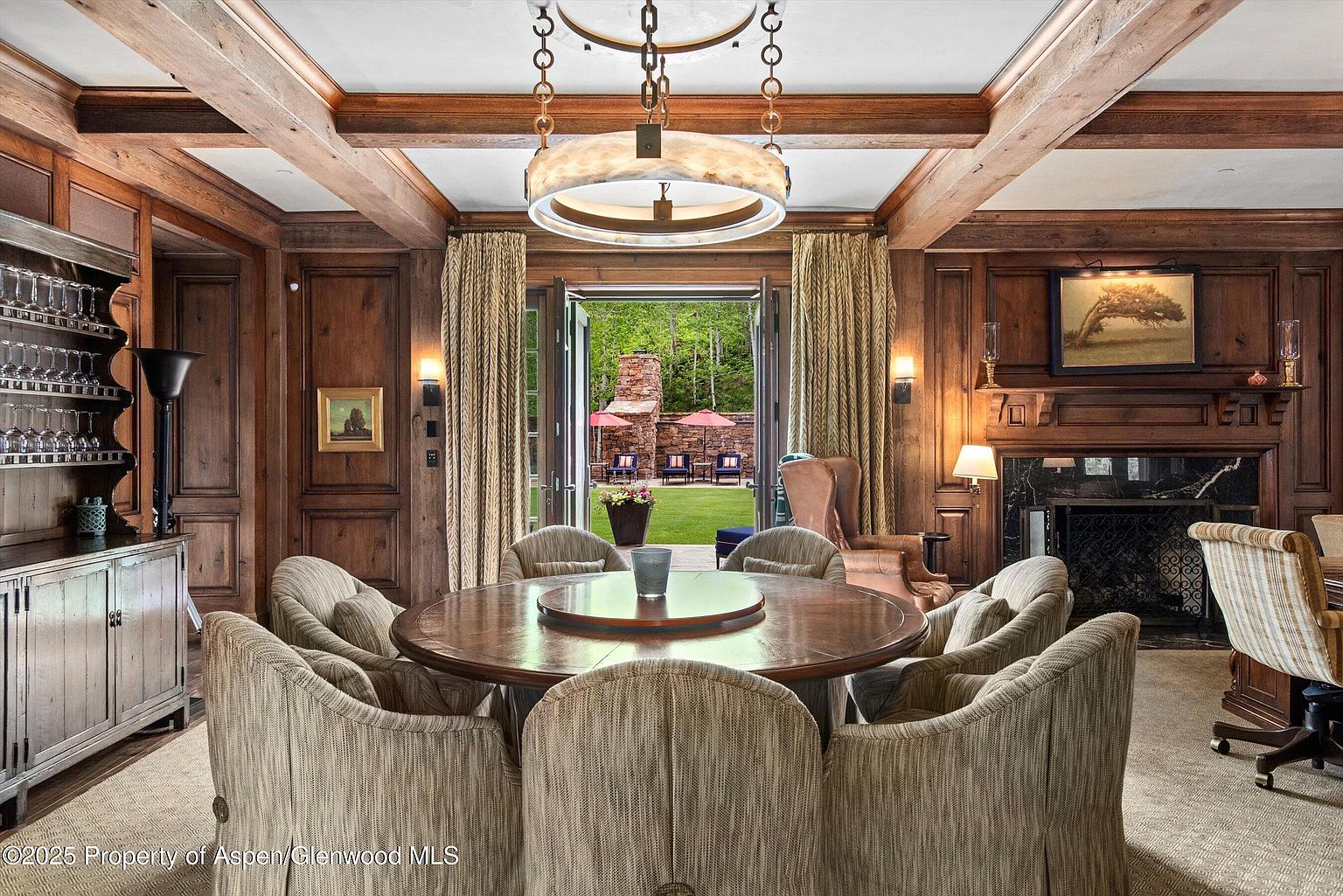 This is an elegant dining room featuring rich wood paneling, a round wooden table surrounded by upholstered chairs, and a striking circular chandelier. French doors open to a view of a well-manicured lawn and outdoor seating area, creating a seamless indoor-outdoor transition. A fireplace adds warmth and character to the space, enhancing the room's luxurious and inviting atmosphere.