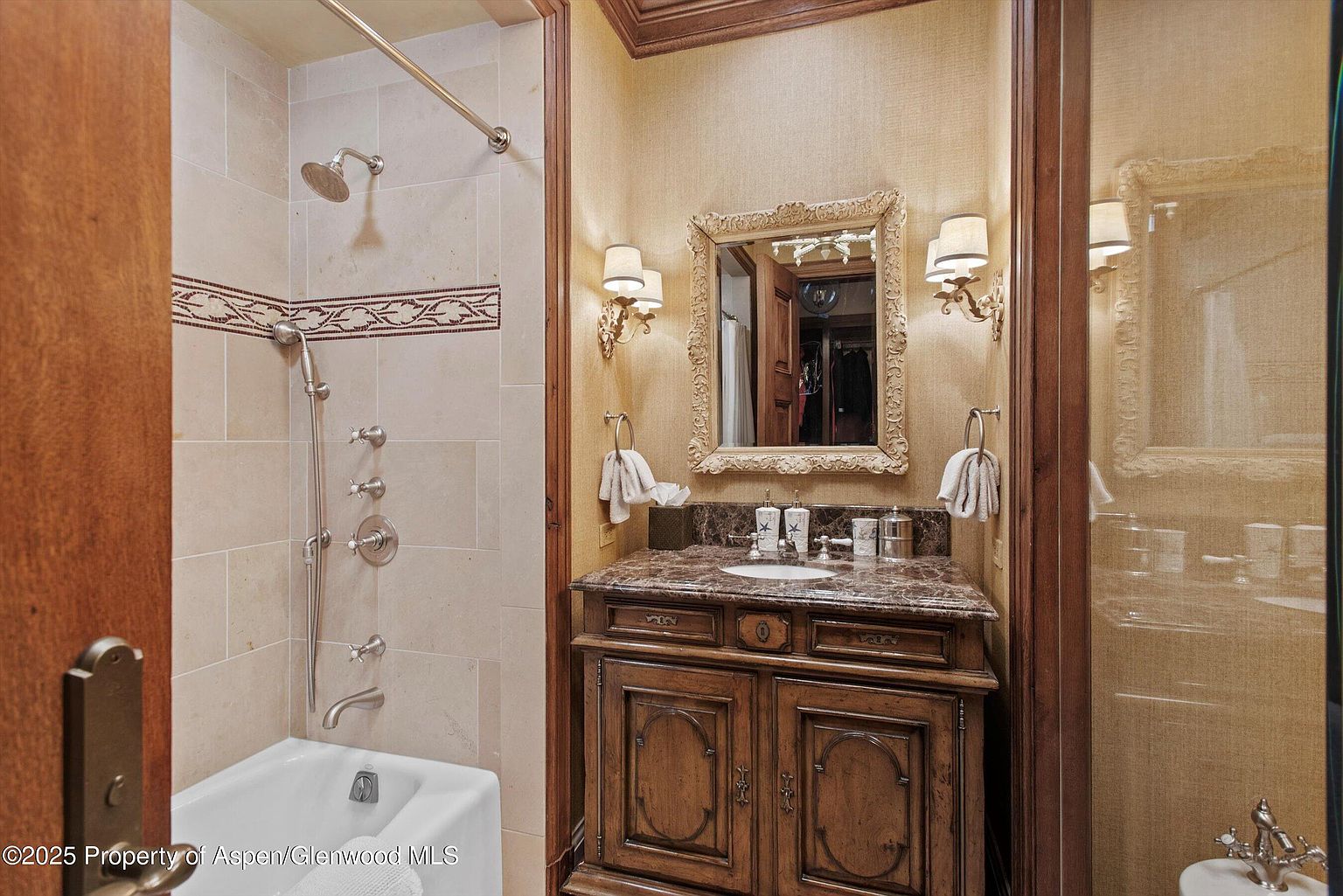 This is a well-appointed guest bathroom featuring a classic design. The vanity is made of dark wood with a marble countertop, complemented by an ornate mirror and sconces. A shower-tub combination is present, enclosed by a glass door, and the walls are covered in a textured wallpaper, creating a warm and inviting atmosphere.