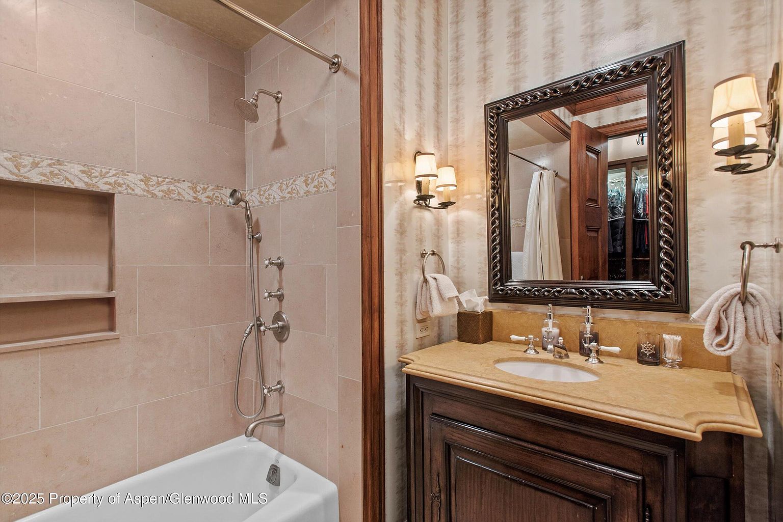 This is a primary bathroom featuring a bathtub with a shower head, a vanity with a marble countertop, and a decorative mirror. The walls are tiled and wallpapered, and the overall style is traditional and luxurious. The perspective is from the doorway, showing the bathtub on the left and the vanity on the right.