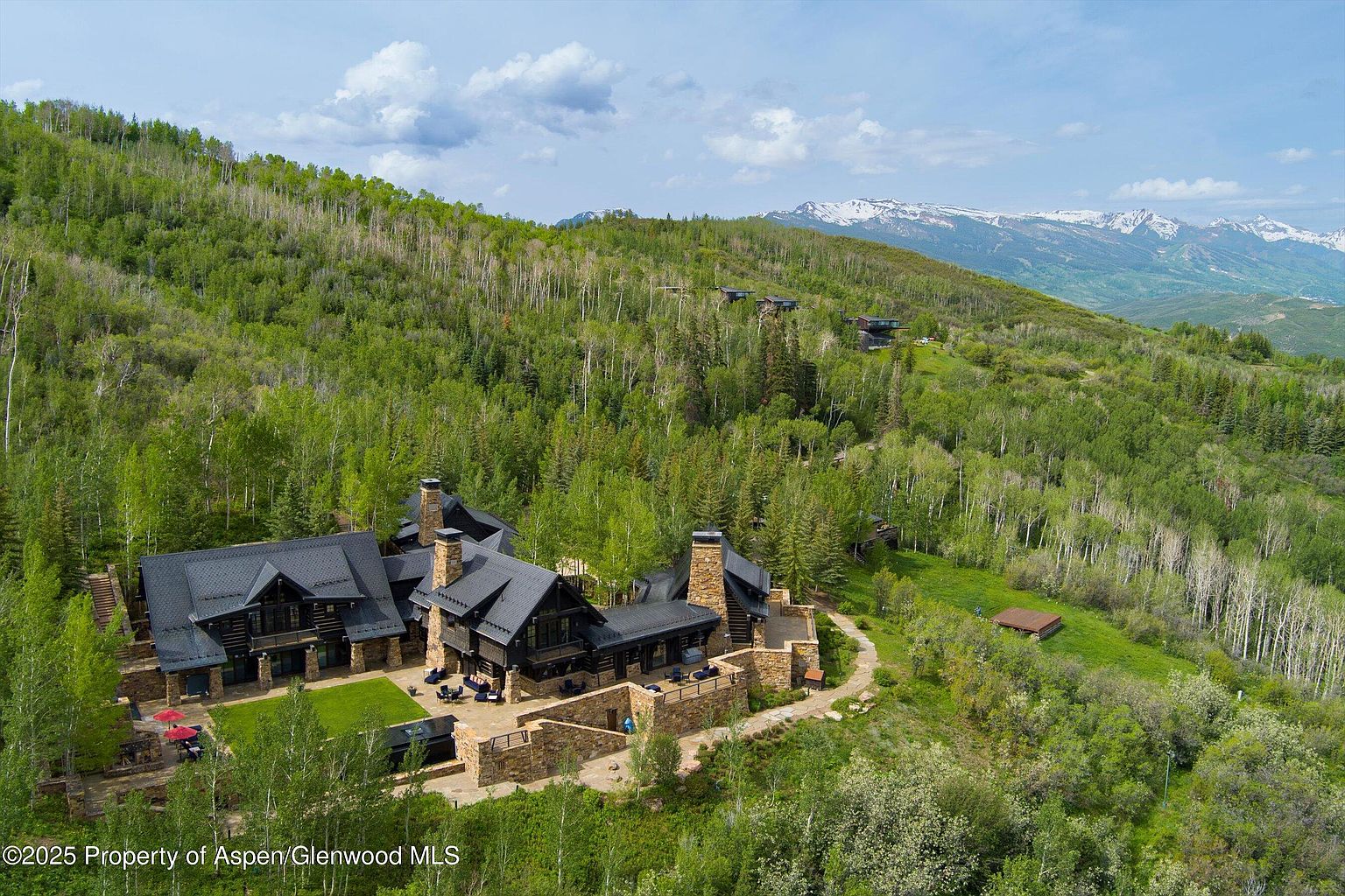 This aerial view showcases a luxurious mountain estate nestled amidst lush greenery. The property features a sprawling residence with dark roofing, stone accents, and multiple outdoor living spaces, including patios and a well-manicured lawn. The surrounding landscape includes dense forests and distant snow-capped mountains, creating a serene and private setting.