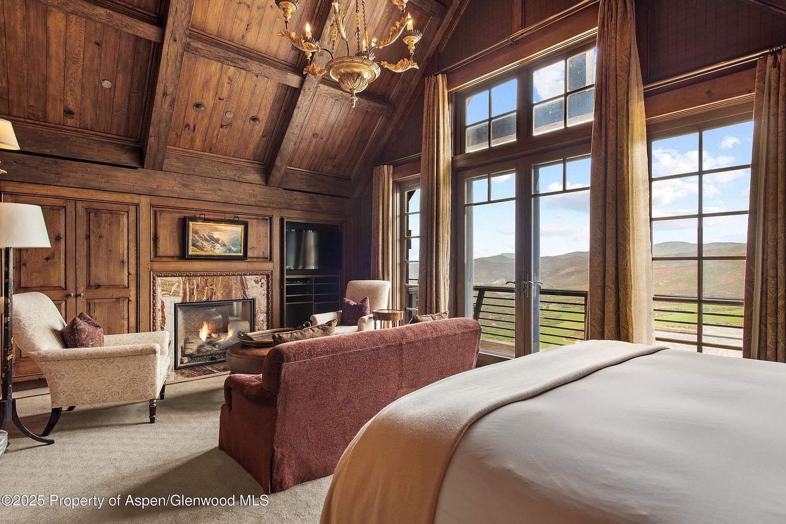 This is a luxurious primary bedroom featuring a rustic, lodge-style design with wood-paneled walls and a vaulted ceiling. A stone fireplace adds warmth, while large windows offer stunning views of the landscape. The room is furnished with a comfortable bed, a sofa, and an armchair, creating a cozy and inviting atmosphere.