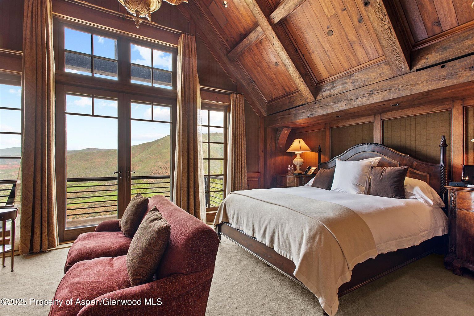 This is a luxurious primary bedroom featuring a rustic, lodge-style design with exposed wooden beams and paneling. A large bed with plush bedding is the focal point, complemented by a comfortable red sofa near the windows that offer scenic mountain views. The room exudes warmth and sophistication, perfect for a high-end real estate listing.