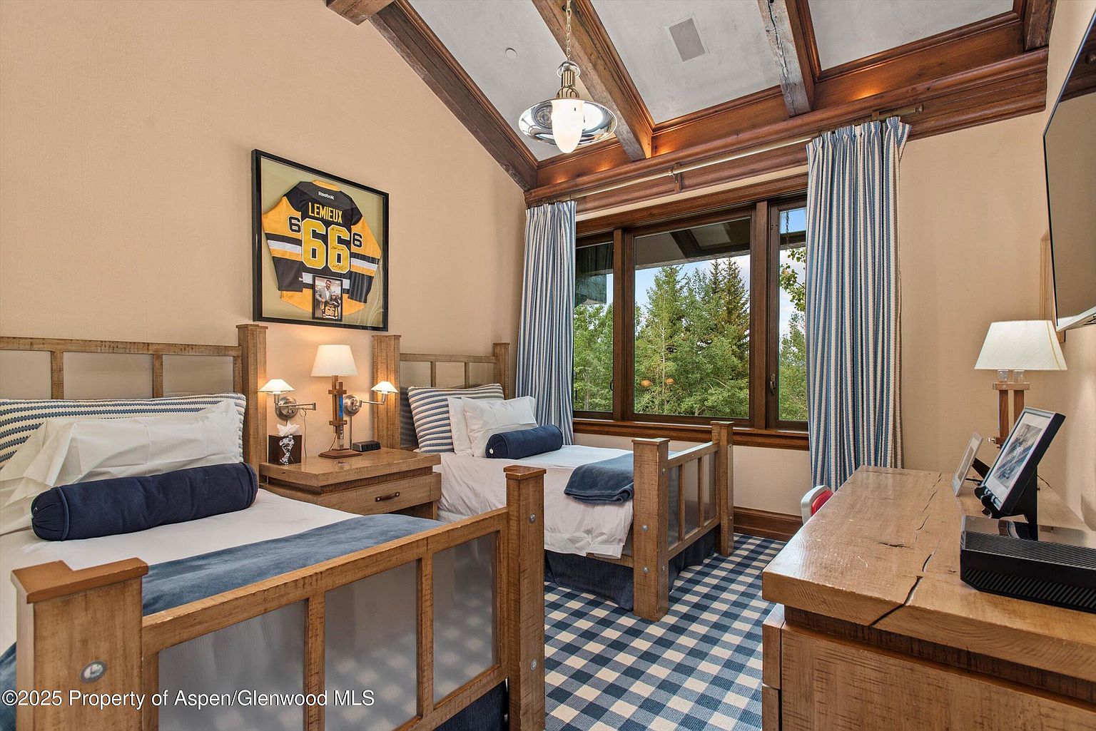 This is a cozy guest bedroom featuring two twin beds with wooden frames and blue and white striped bedding. A framed sports jersey hangs above one of the beds, adding a personal touch. The room has a rustic-chic style with exposed wooden beams on the ceiling and a blue and white checkered carpet, creating a warm and inviting atmosphere.