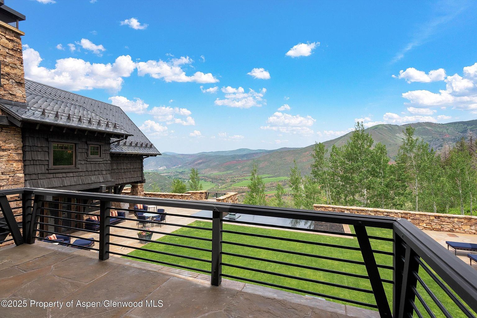 This image showcases a luxurious patio or balcony with a stunning mountain view. The space features a stone floor, a black metal railing, and outdoor furniture, suggesting a comfortable area for relaxation and entertainment. Beyond the patio, a well-maintained lawn and a reflective pool add to the property's appeal, while the backdrop of rolling hills and a clear sky enhances the sense of tranquility and exclusivity.
