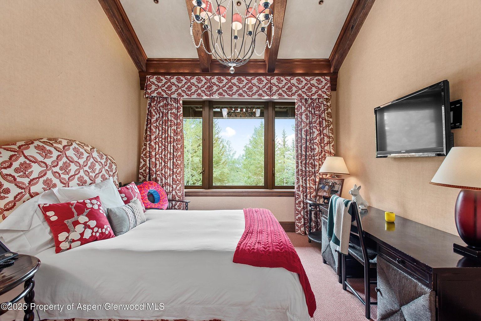 This is a cozy bedroom featuring a bed with patterned headboard and decorative pillows, a wooden desk with a chair, and a wall-mounted television. The room has a warm color palette with patterned curtains and a chandelier, creating a comfortable and inviting atmosphere. The window offers a view of the trees outside.