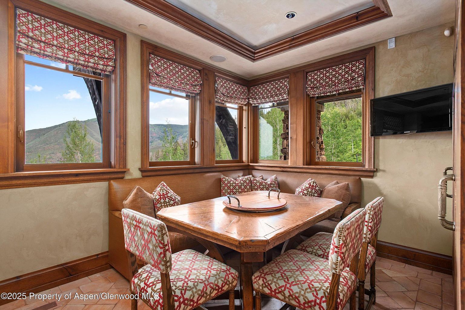 This dining room features a cozy breakfast nook with built-in seating and a wooden table, perfect for intimate meals. The room is surrounded by windows offering scenic views of the outdoors, complemented by patterned window treatments and chair upholstery. A mounted television adds a modern touch to the rustic, wood-paneled aesthetic.