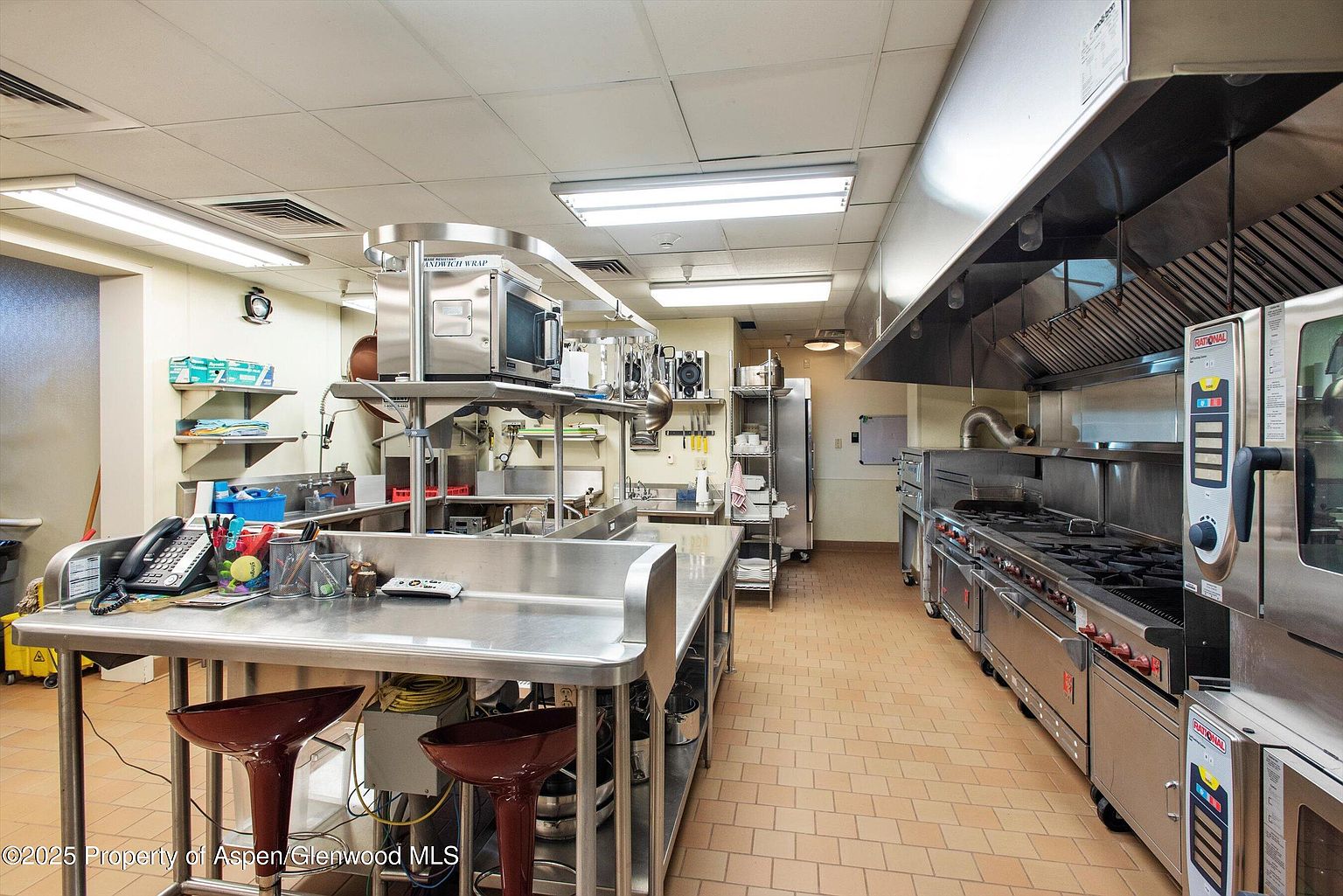 This is a commercial kitchen featuring stainless steel countertops, appliances, and shelving. The space is well-lit with fluorescent lighting and has a tiled floor. There are multiple workstations and cooking areas, including a large range and oven.