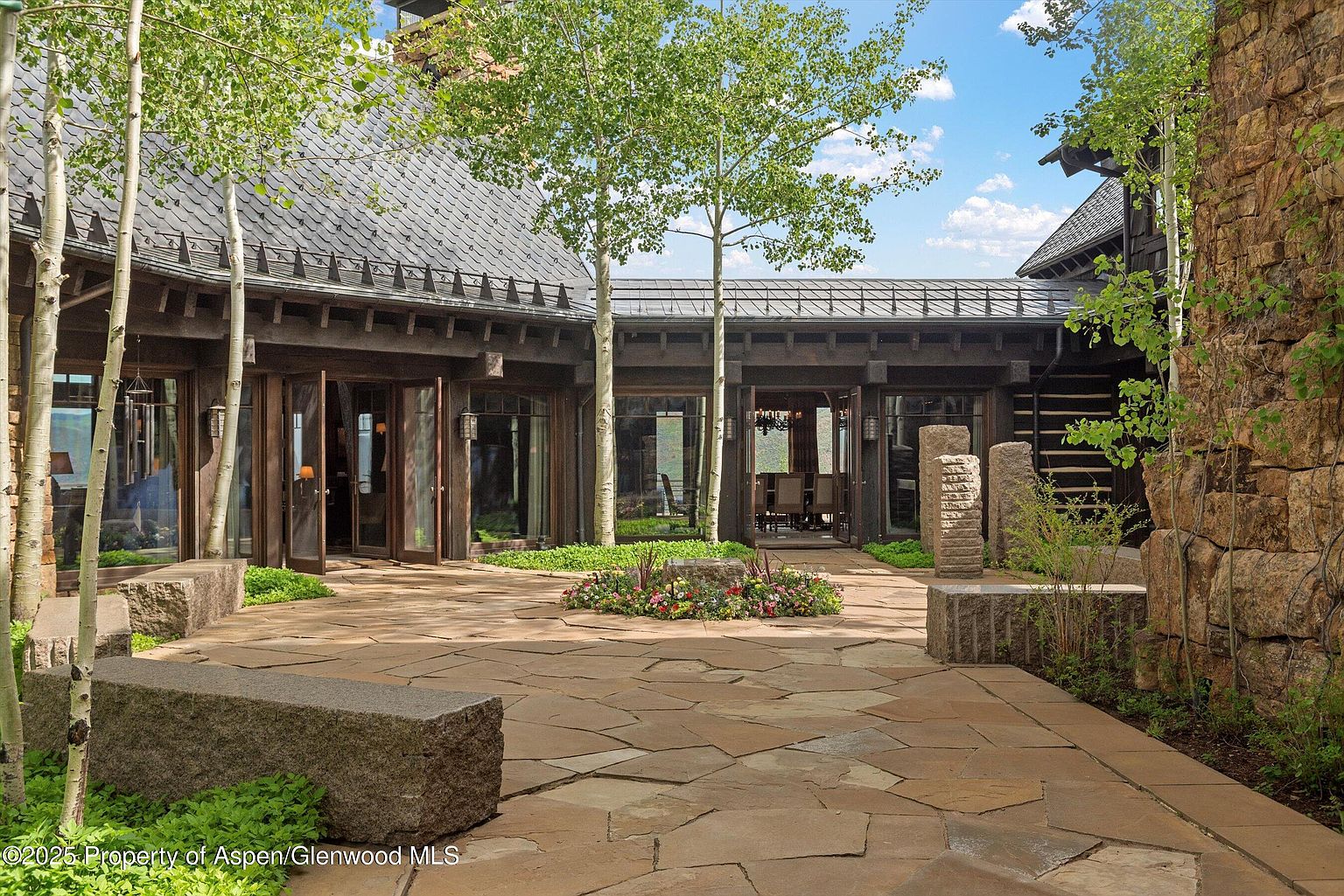 This image showcases a beautifully designed outdoor patio area. The patio features a stone-paved surface, complemented by stone benches and landscaping with trees and flower beds. The architecture of the surrounding building is rustic and elegant, with dark wood and large glass doors that open to the interior, creating a seamless indoor-outdoor living experience.